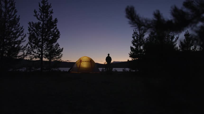 Man standing near lighted tent at night looking out to lake while camping