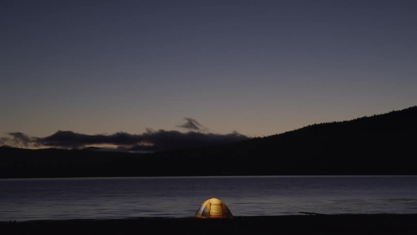 A tent being illuminated by a light within on the shore of a lake
