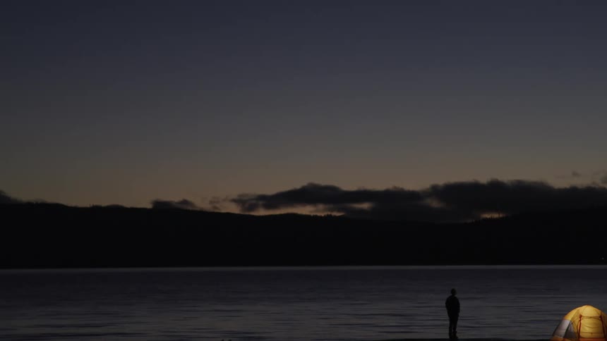 A man stands by his tent and looks out at the lake