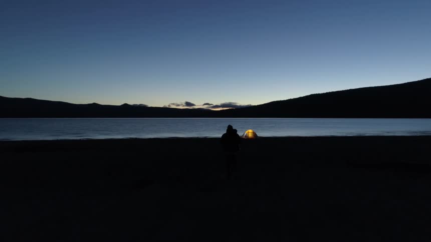 A person walks away from a lakeside campsite and tent at night