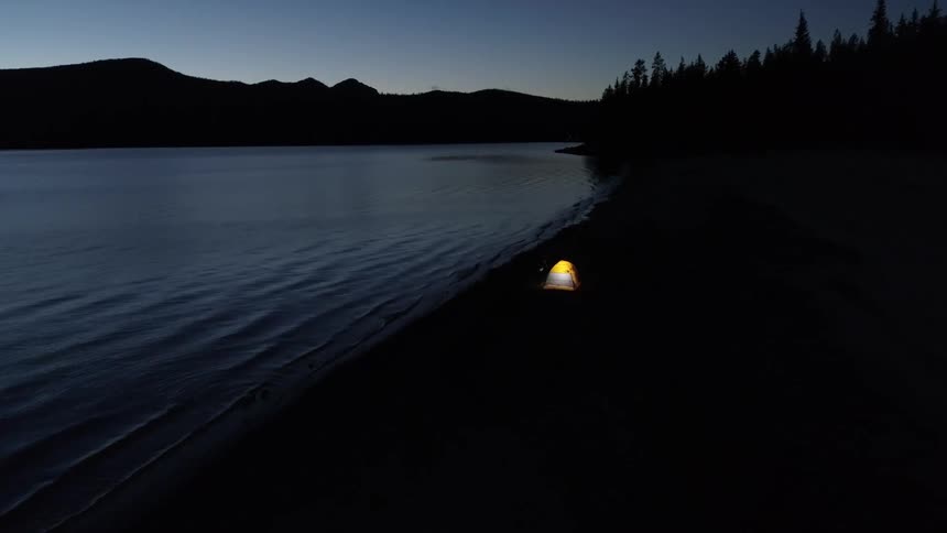 A lit up tent is set up next to the lake in a forest near a mountain range