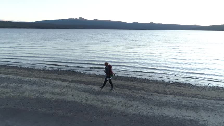A person walks along the shoreline with a backpack and sleeping bag