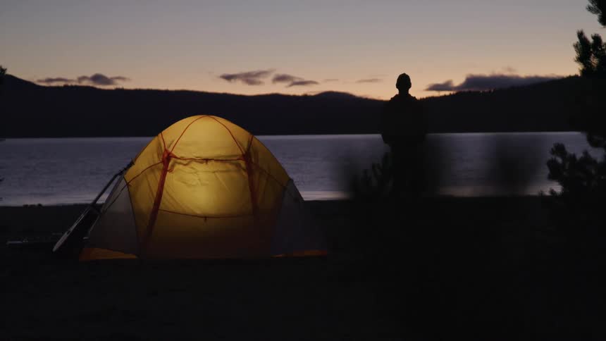 A person stands beside a lake with a lighted tent behind them