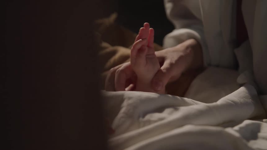 A woman holds a babies hand at they lie in bed