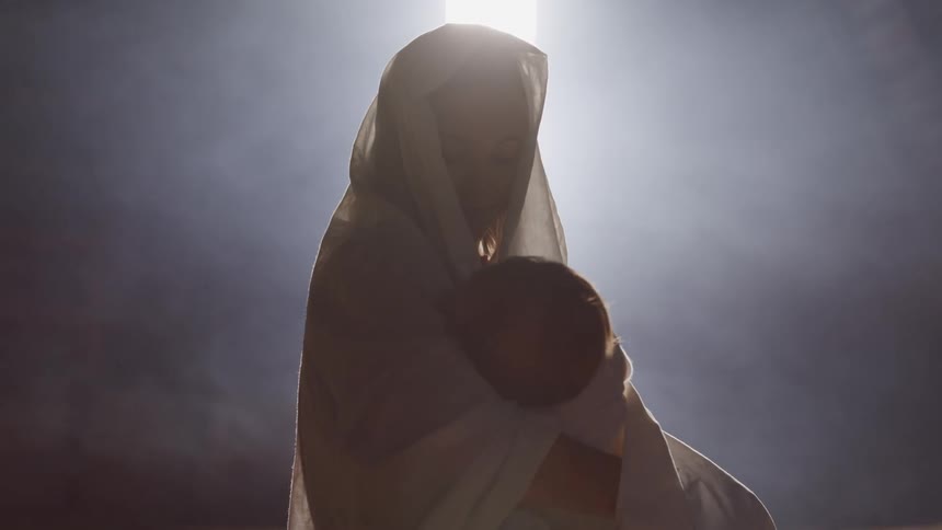 A woman in a shawl holding a child while sitting in a barn illuminated by sunlight