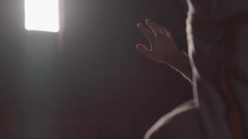 A man with a scarf covering his head reaches out toward a small window