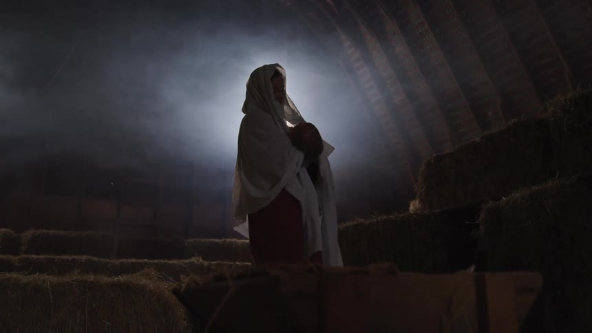 A woman in a shaw rocking a baby while standing in a barn filled with hale bales
