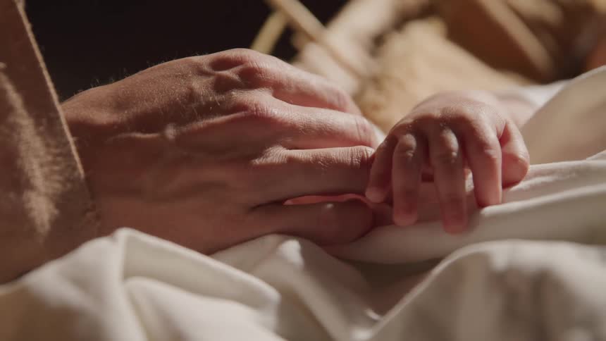 An adult resting their hand next to the hand of a baby lying in a manger