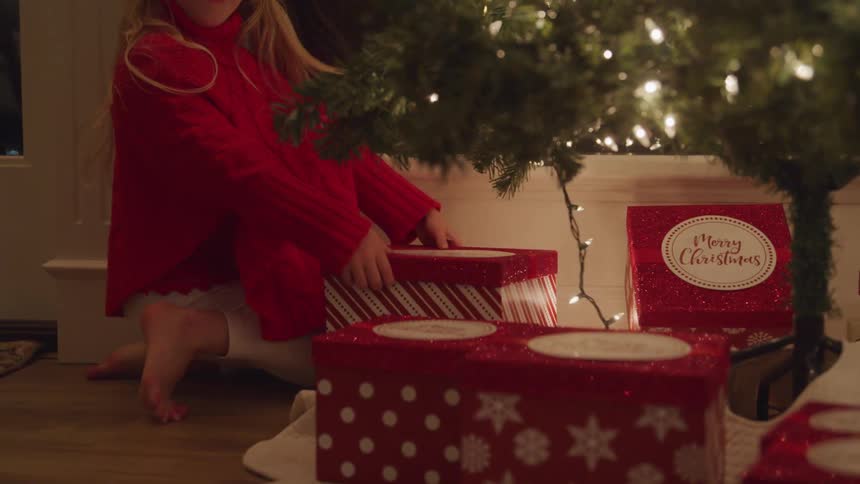Female child runs to a present placed under a lit christmas tree shakes the box and then removes the lid from the box and finds a string of balls