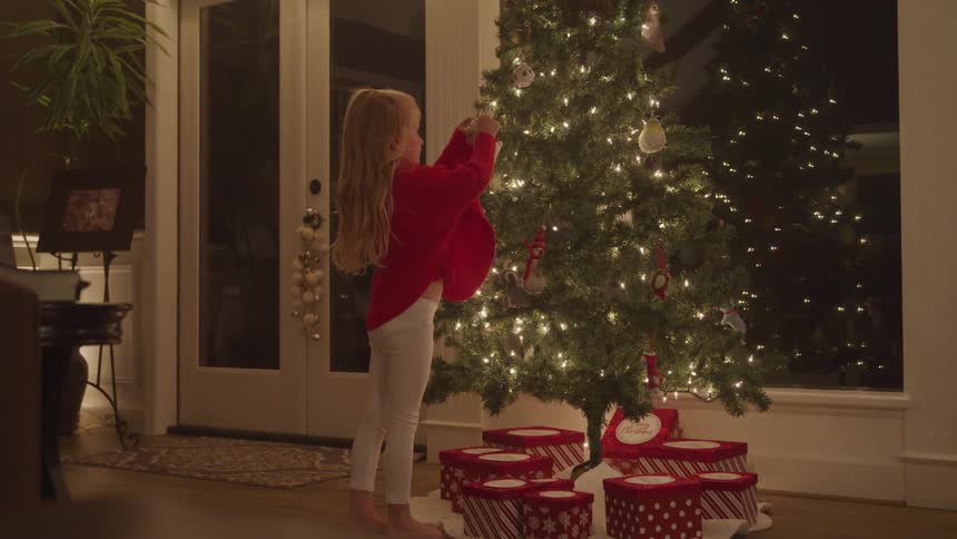 Female child walks up and hangs an ornament on a christmas tree with the lights on and presents carefully arranged underneath after dark