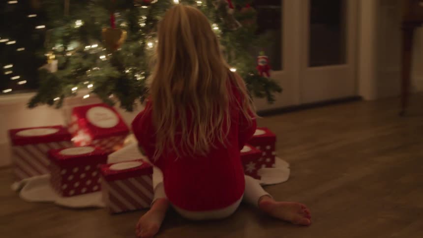 A young girl runs to a christmas tree and picks up a gift