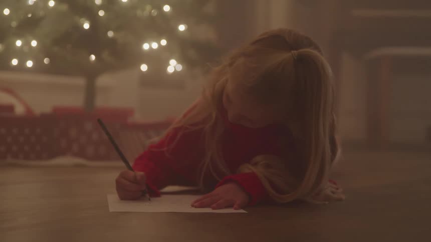 A young girl lying on the floor before a christmas tree writing on a sheet of paper