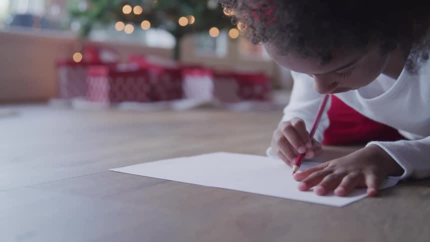 Black child writes a letter to santa in front of the christmas tree in the living room at home