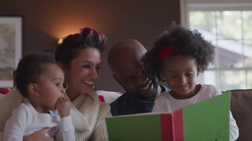 Mother and father along with their two children are sitting on the couch together reading a book and laughing together