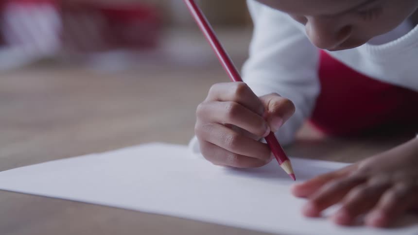 Child kneels on floor drawing on white paper with red pencil