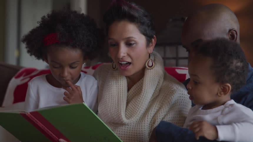 A woman reads a book to her daughters with her husband beside