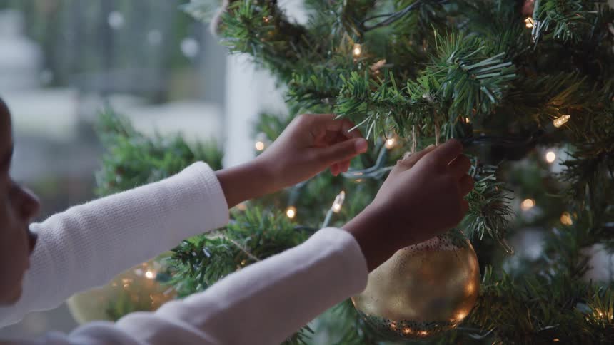 Black child decorates christmas tree with christmas bulbs in the living room at home