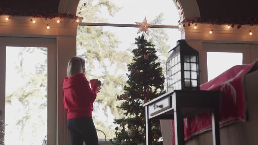 A woman with a teacup walking up to a christmas tree than sipping on tea