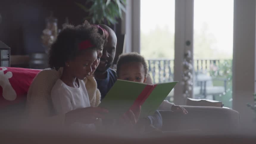 Black parent reads and leafs through a story book for their children in the living room at home