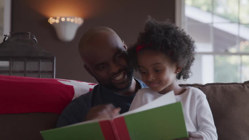 Black father and daughter sitting on the sofa read a story book together in the living room