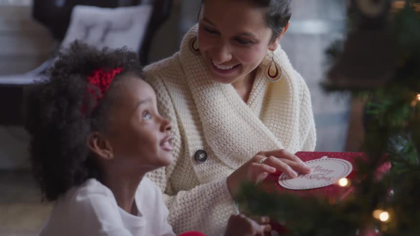 Woman and child sitting next to the christmas tree try to guess what is inside the gift box in the living room