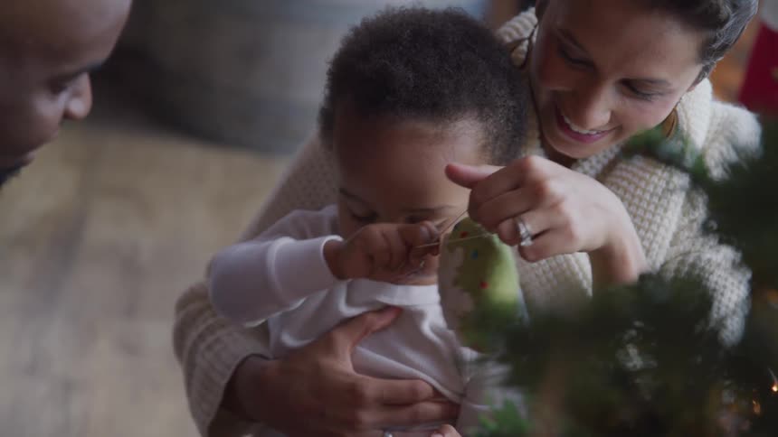 A dad looks on smiling as his wife holds their son and plays with the christmas ornament he is holding