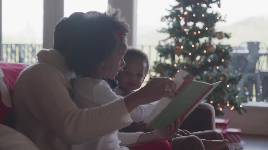 Mother and daughters sitting on the couch read a book near the christmas tree