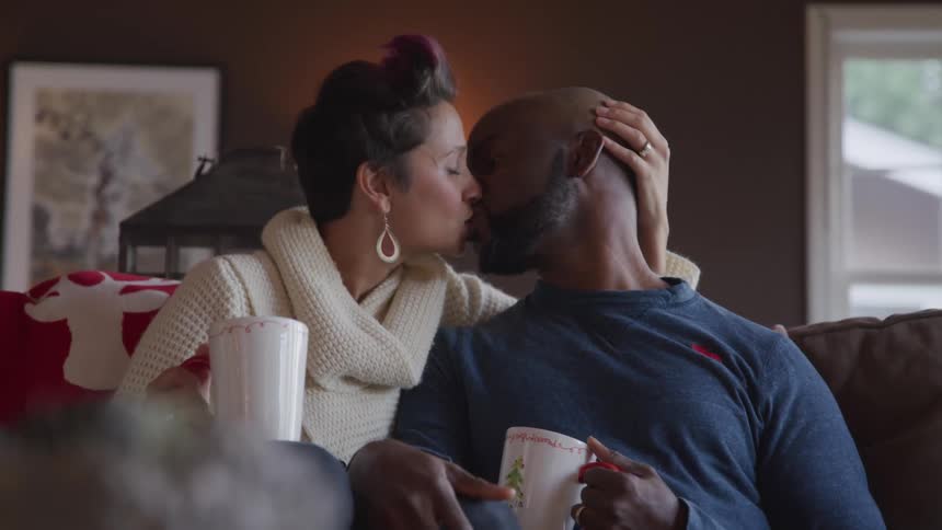 White woman and black man sitting on the couch kiss each other in the living room at home