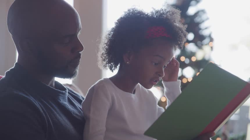 Father and daughter sitting on the sofa read a story book next to the christmas tree