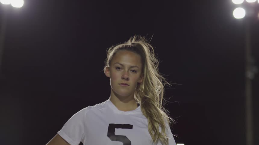 Blonde female footballer stands and looks at the camera on a football field at night