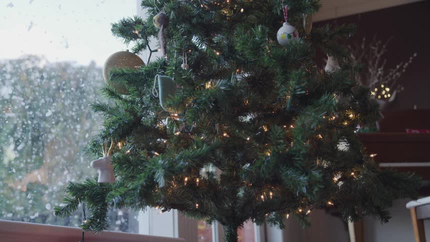 A christmas tree covered in lights and decorations next to a window through snow can be seen falling on the other side