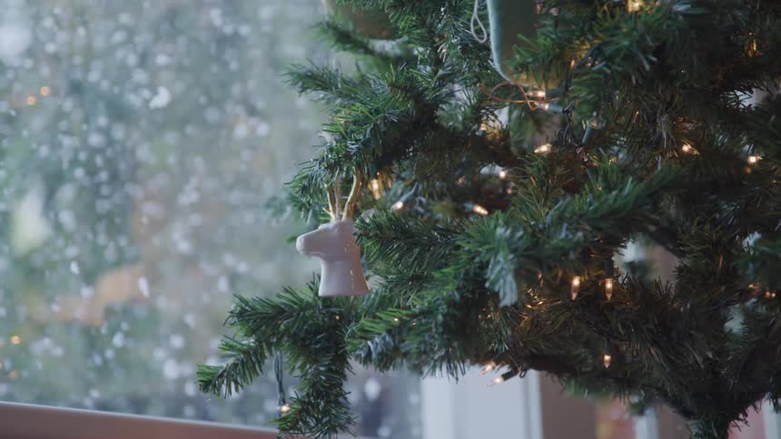 Christmas tree decorated in white with lights stands near window while it snows