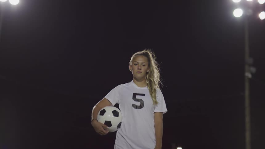 Female footballer holding a ball stands and looks at the camera on the football field at night