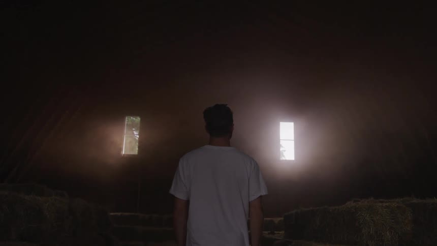 Man stands in barn near stacked hay while sunlight shines through two high windows