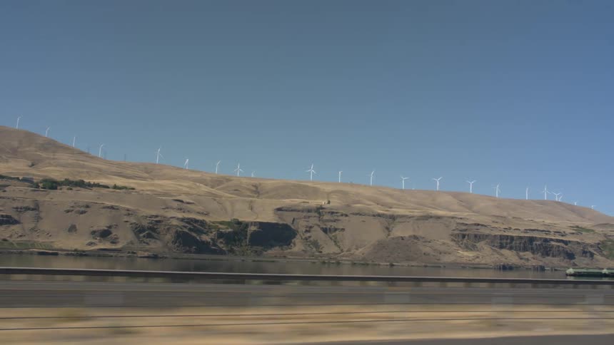 Wind turbines sit on top of a sand hill that runs alongside a river