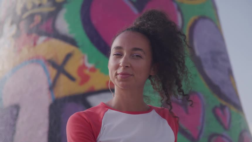 A young woman turning to smile into the camera while standing in front of a column covered in colorful street art