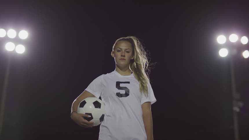 Female footballer stands and hold a ball on the football field at night