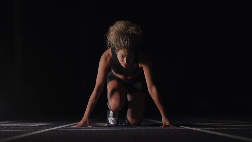 A female athlete kneeling in preparation for a sprint on a running track in the darkness
