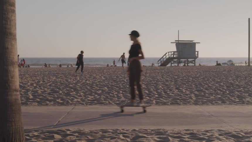 Woman skating and men riding bikes in a sidewalk in front of a beach at dusk