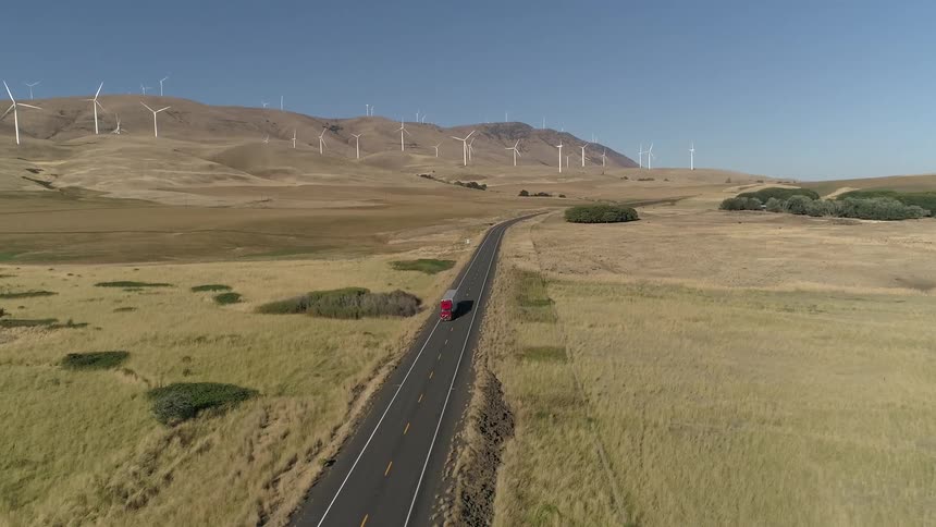 Semi with red cab drives along road away from wind farm occupying hills