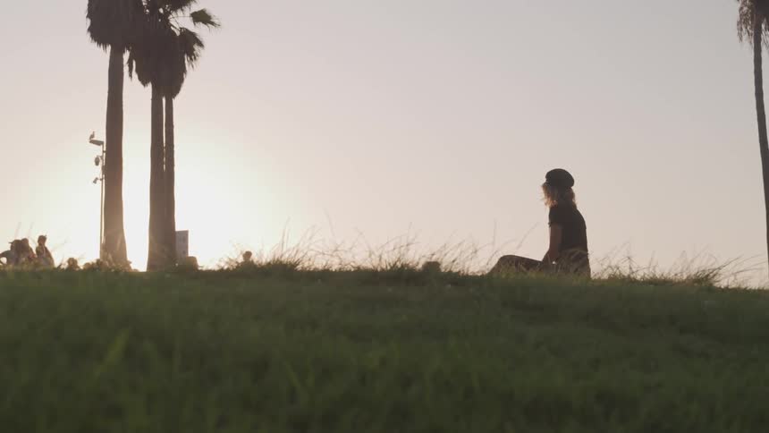 Female sitting at the top of a grassy hill watching people move past during as the sun is beginning to set