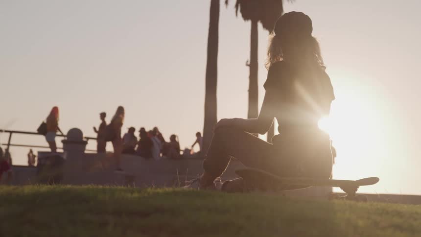 A woman sitting on a skateboard with people trees and the sun in the foreground