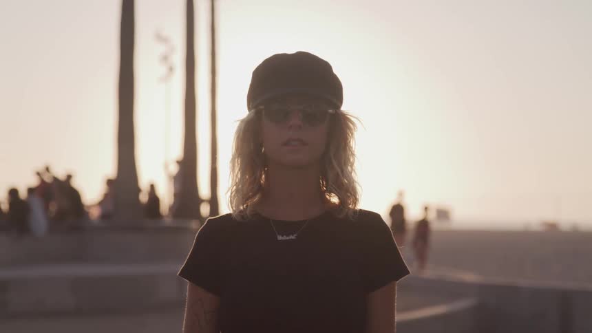 Blonde woman wearing sunglasses and a black hat poses for the camera outdoors at sunset