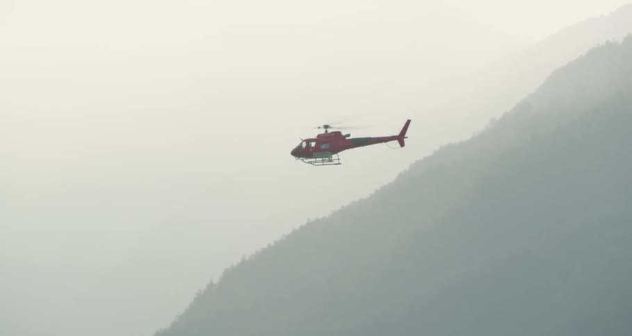 Red helicopter flies near the mountains under a cloudy sky