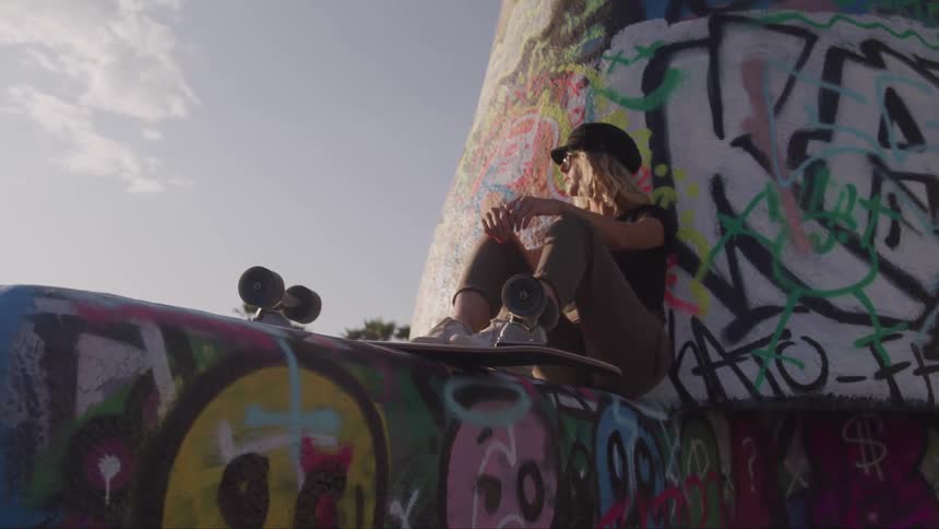 A woman sits outdoors against a graffitied wall with a skateboard under her feet