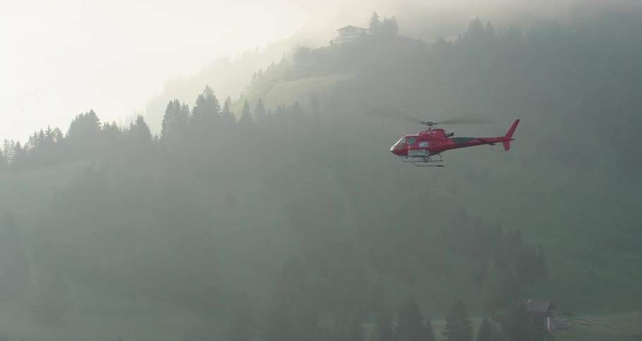 A red helicopter flying over a forest shrouded in fog