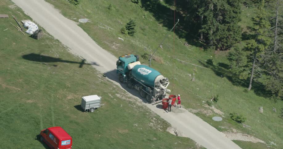 Two men guiding a large barrel by a cement truck with the shadow of a helicopter above