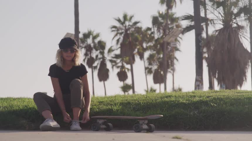 Female tying her shoes then picking up skateboard and walking away at the beach