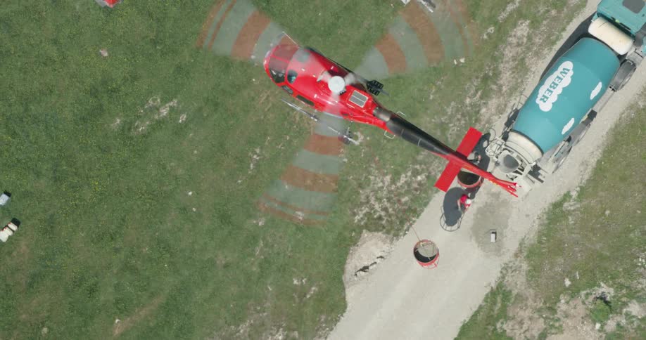 An aerial view of a hovering helicopter lifting away a large bucket of cement