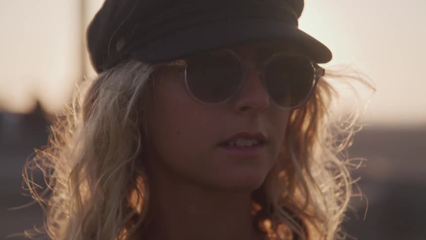 Blonde woman wearing sunglasses and a hat poses for the camera at sunset